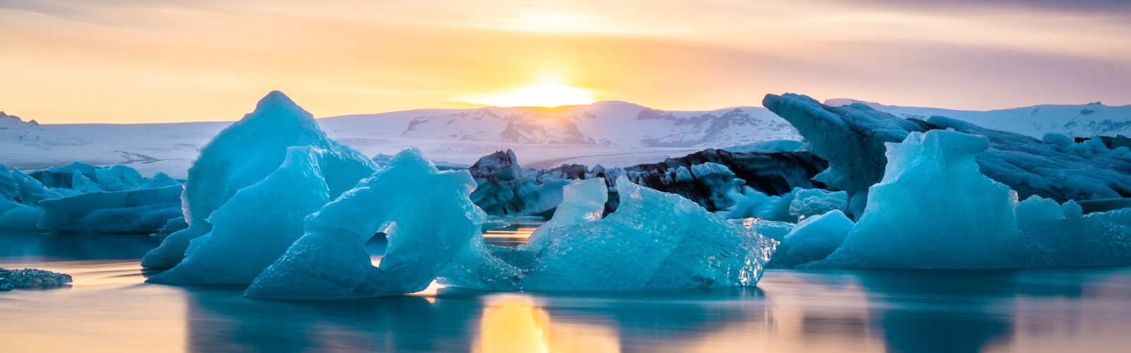 The rising sun is just visible over snowy mountains. In the foreground, there are five icebergs floating in the water. 